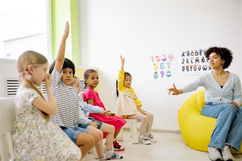 kids sitting in a circle raising hand with teacher in front of them 